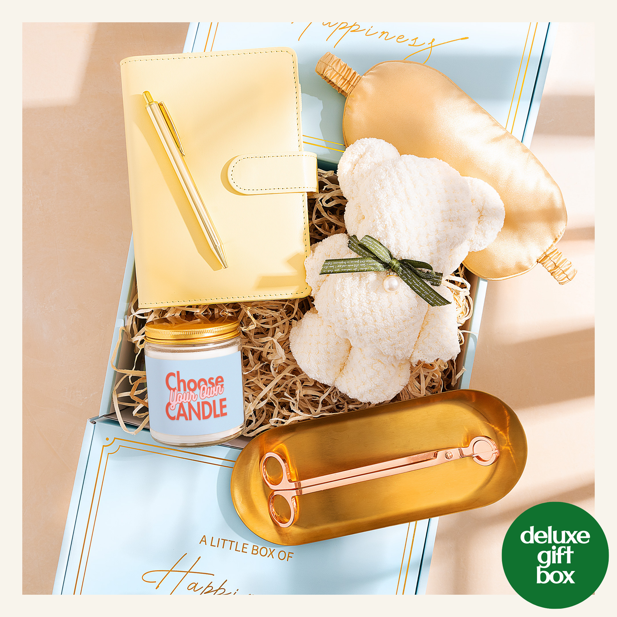 A gift box containing a variety of items, including a yellow notebook, a white teddy bear, a gold candle, a gold tray, and a gold pair of scissors.